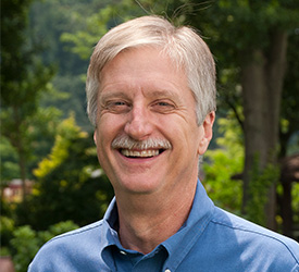 Man smiles in front of trees and shrubs.